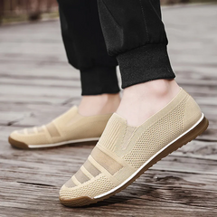 A person wearing lightweight slip-on mesh shoes with a textured design, cushioned insole, and brown soles stands on wooden planks outdoors. The person is dressed in black jogger pants.