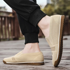 A person wearing lightweight slip-on mesh shoes with brown soles, standing outdoors on a wooden surface, dressed in black cuffed pants.
