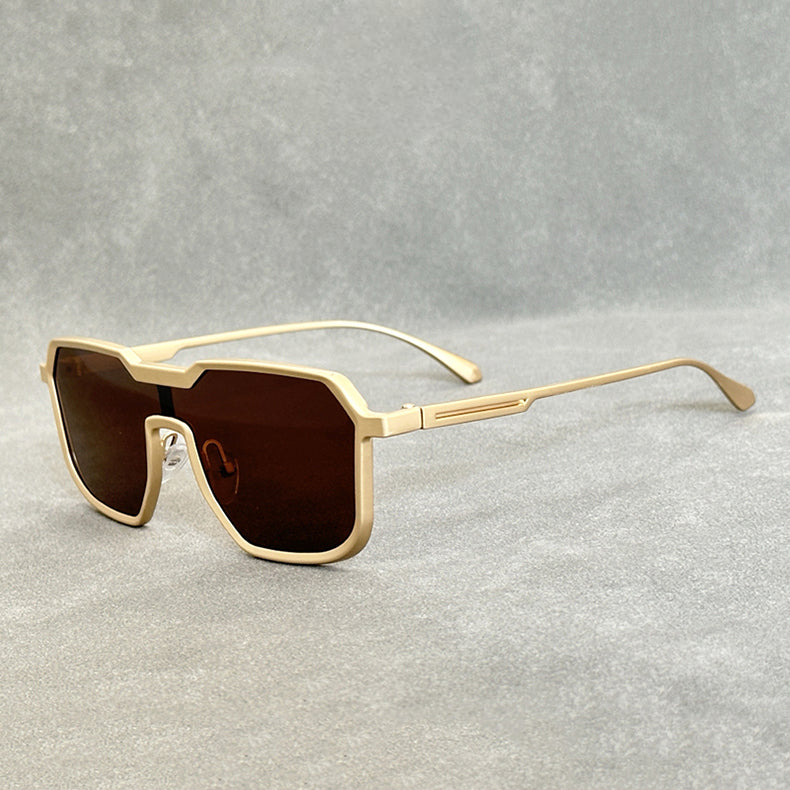 Beige, geometric square-frame sunglasses with dark brown lenses are shown on a plain, light gray surface. The design features angular edges and a modern, oversized style inspired by steampunk sunglasses.
