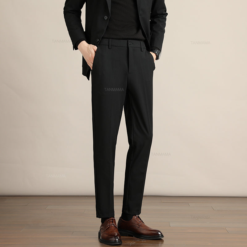 A person wearing a black suit with a black shirt and Mens Slim Casual Trousers stands on a wooden floor against a plain beige background. Brown leather dress shoes complete this business formal look. The persons head is not visible in the image.
