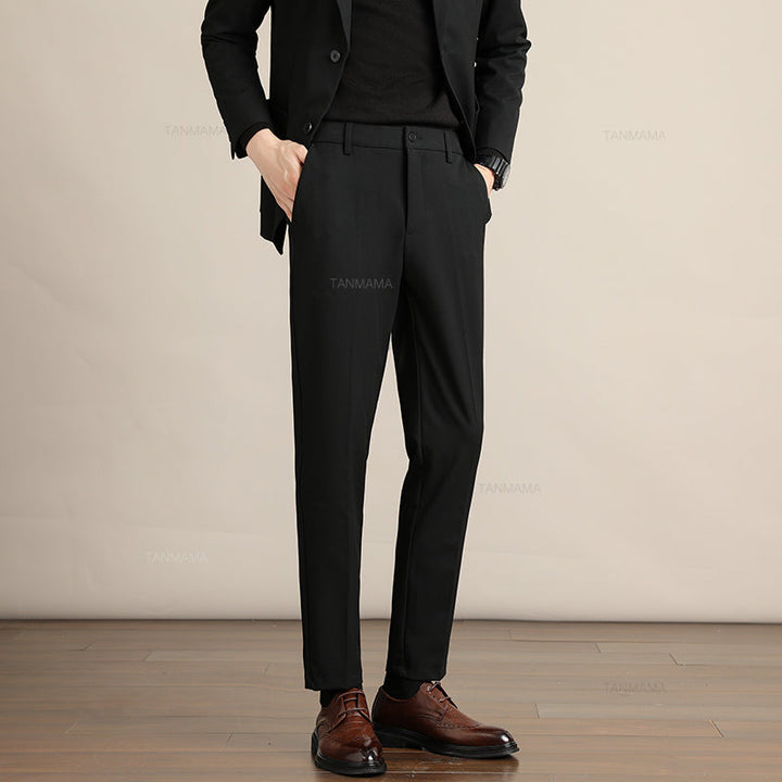 A person wearing a black suit with a black shirt and Mens Slim Casual Trousers stands on a wooden floor against a plain beige background. Brown leather dress shoes complete this business formal look. The persons head is not visible in the image.