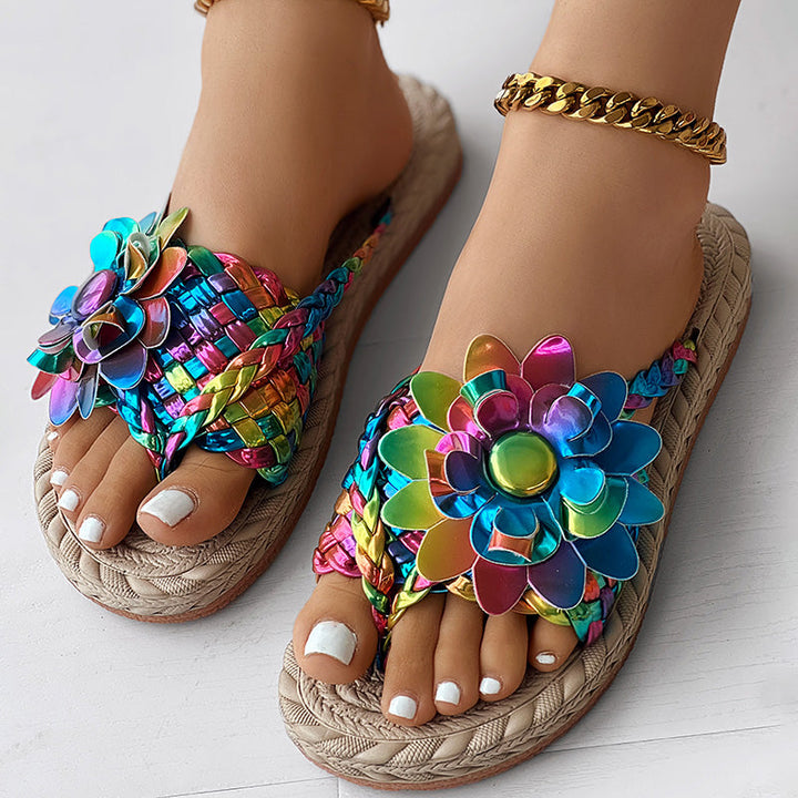 A person wearing colorful, woven sandals decorated with large, shiny, rainbow-colored flower designs—a vacation essential in women's fashion casual shoes—with a gold anklet on one ankle and white-painted toenails.