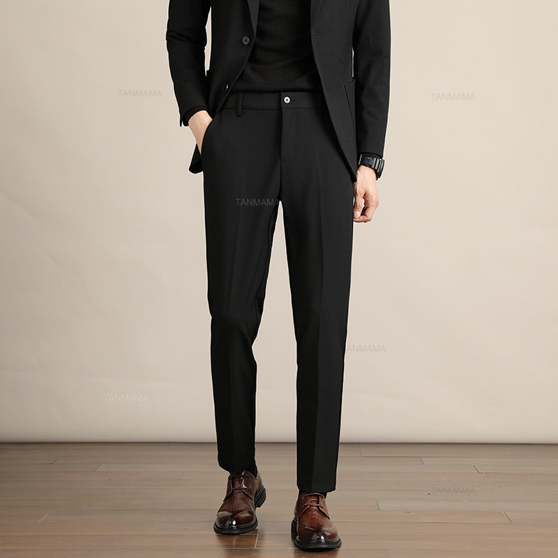 A person wearing a black suit, black shirt, and brown dress shoes stands with one hand in their pocket against a beige background on a wooden floor, showcasing Mens Slim Casual Trousers crafted from premium fabric. The persons face is not visible.