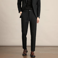A person wearing a black suit, black shirt, and brown dress shoes stands with one hand in their pocket against a beige background on a wooden floor, showcasing Mens Slim Casual Trousers crafted from premium fabric. The persons face is not visible.