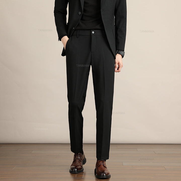 A person wearing a black suit, black shirt, and brown dress shoes stands with one hand in their pocket against a beige background on a wooden floor, showcasing Mens Slim Casual Trousers crafted from premium fabric. The persons face is not visible.