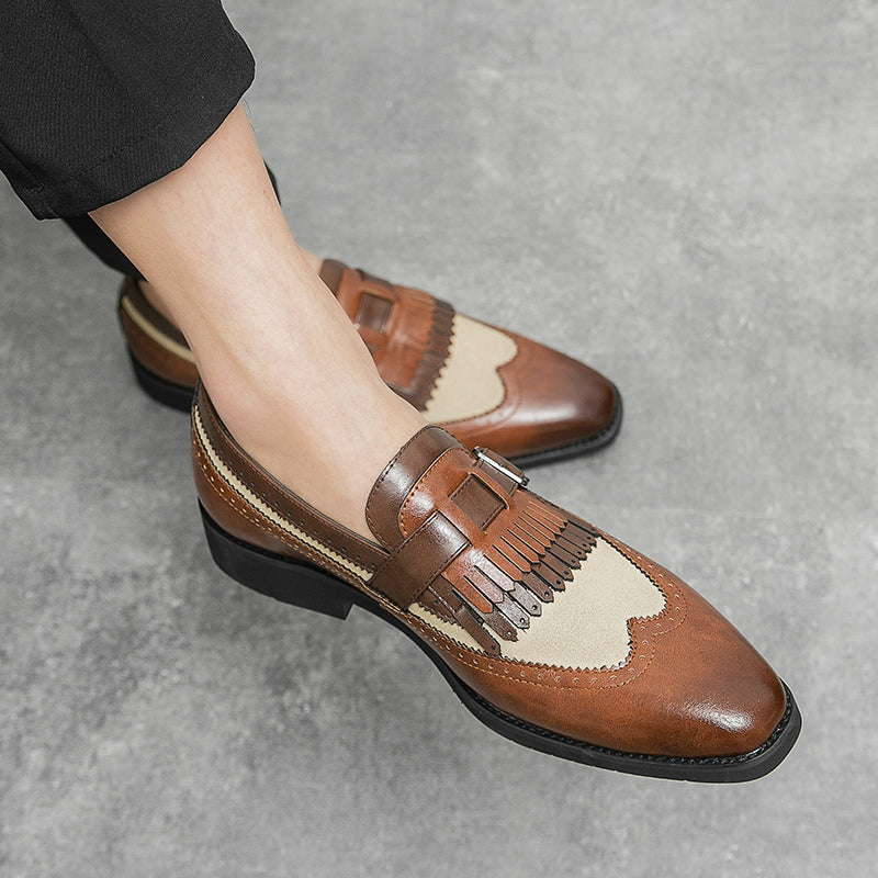 A person wearing stylish brown and beige premium leather shoes with fringe detailing and buckle accents, featuring a slip-on design, stands on a gray textured floor.