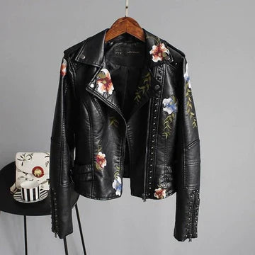 An embroidered moto jacket in black PU leather with studded details and colorful floral embroidery hangs on a wooden hanger against a gray wall. A small table with floral-patterned teacups sits nearby.