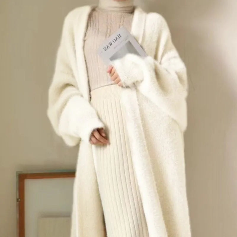 A person wearing a long, cream-colored, cozy cardigan with a relaxed drape fit over a beige turtleneck and ribbed skirt holds a book against their chest. The outfit has a soft, elegant look in neutral tones. The background is minimal.