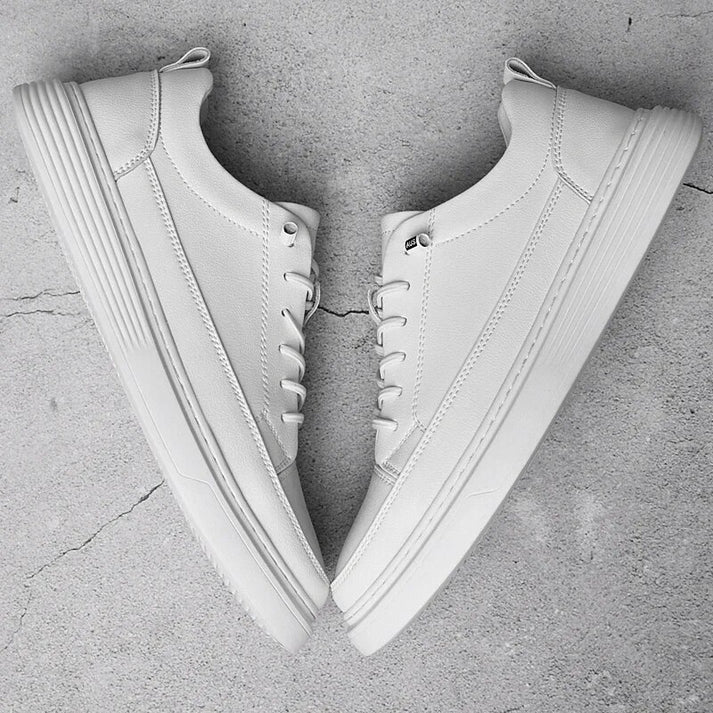 A pair of clean, white low-top sneakers with laces—crafted from genuine leather—are placed side by side on a cracked concrete surface, forming a mirror image of each other.